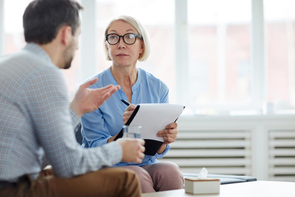 Blonde mature woman making notes in document while looking at patient and listening to his story